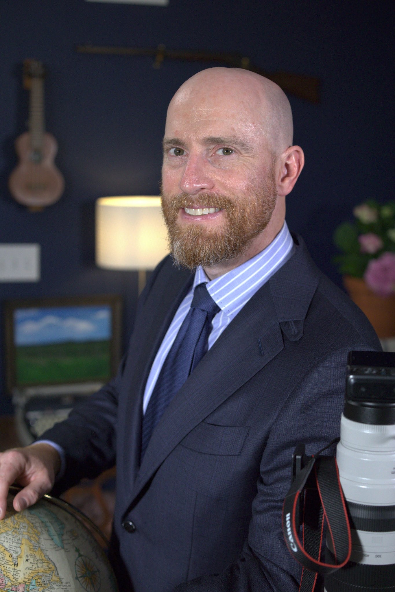 Charles Edward Pax smiling indoors, with a camera, globe, and other objects nearby